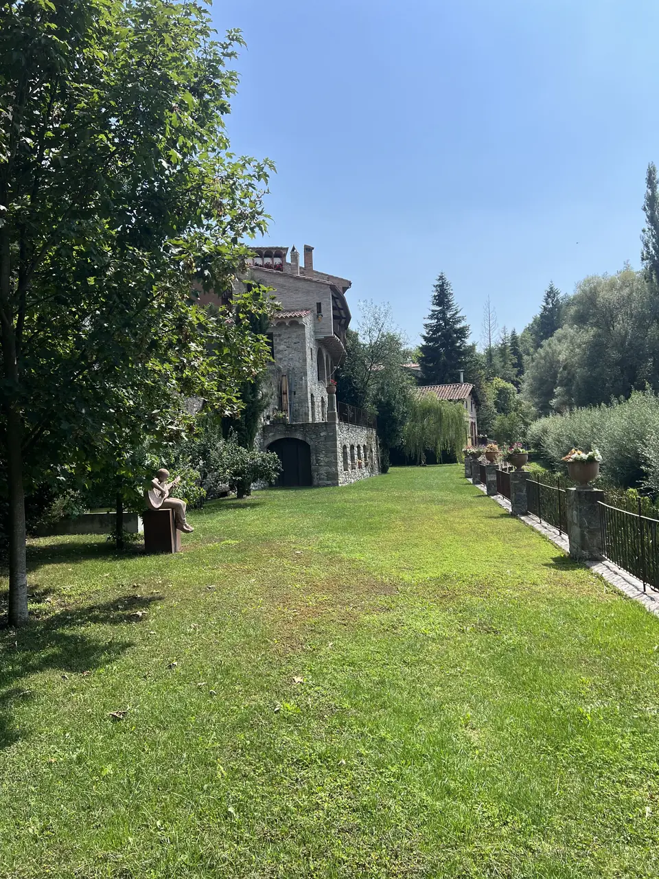 Jardín alargado de césped junto a una casa de piedra, con árboles y valla de madera